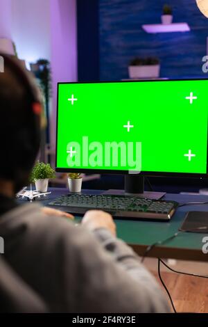 Gamer man playing videogames on computer with green screen holding wireless joystick in professional gaming studio. Player using pc with mock up isolated desktop streaming shooter games wearing headset Stock Photo