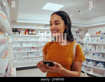Young woman smiling while using smartphone to read prescription and chose medicine from shelf in pharmacy Stock Photo