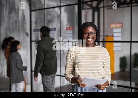 Waist up portrait of young African-American businesswoman standing by glass wall while planning project in modern office, copy space Stock Photo