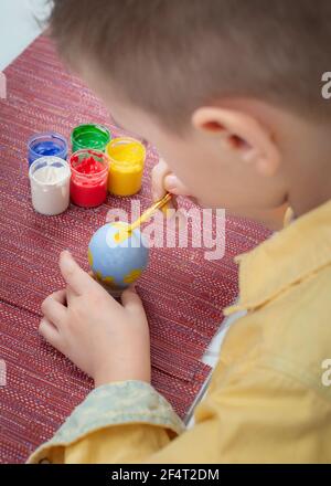 A boy painting an Easter egg, on a red fabric background. Painting Easter eggs, gouache paints, brush, preparation for the holiday Easter. Vertical photo. Focus on the egg Stock Photo