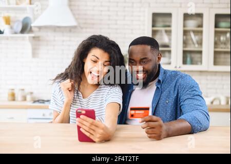 Overjoyed biracial couple shopping together online, excited multiracial woman and man hold smartphone and a credit card, rejoy purchases online, making order, buying something in e-shop Stock Photo