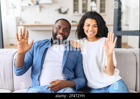 Video screen with cheerful multiracial couple, looks at the camera and waving. Webcam view of smiling young biracial woman and man sitting on the couch and greeting, virtual meeting concept Stock Photo
