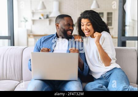 Overjoyed cheerful multiracial couple celebrating good news looking at the laptop screen, young excited woman and man scream happily sitting with computer on the cozy couch at home, won in video game Stock Photo