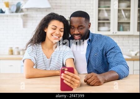 Lovely multiracial couple taking a selfie together sitting at the kitchen desk, smiling biracial woman and cheerful African guy hold smartphone, look at the webcam. Couple is web surfing on the phone Stock Photo