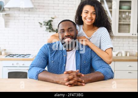 Portrait of African-American couple in love in the kitchen at home, a beautiful biracial woman hugs the shoulders of handsome black man, they look at the camera and smile spending time at home Stock Photo