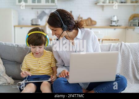 Happy mother with kid sitting on sofa at home during lockdown work on laptop child playing in tablet Stock Photo