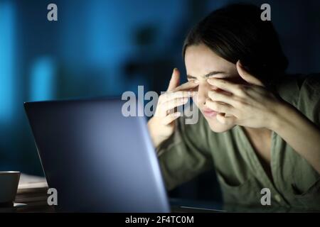 Fatigued woman with eyestrain scratching eyes using laptop late hours in the night at home Stock Photo