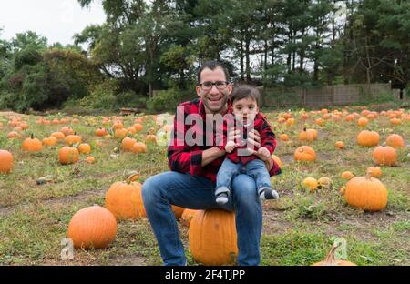 Handsome father and adorable infant son pose for autumn holiday portraits sitting on large pumpkin in a field wearing matching plaid button down flann Stock Photo