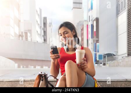 A HAPPY YOUNG WOMAN SITTING AND USING MOBILE PHONE IN A STREET Stock Photo