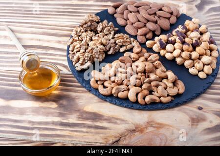 Various nuts sorted in glass bowl with honey bowl and honey dipper. Mixed nuts on wooden table. Black stone plate on wooden background. Stock Photo