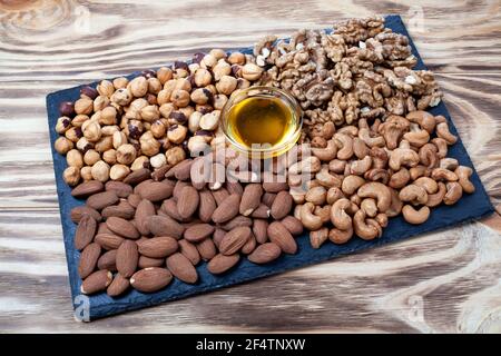 Various nuts sorted in glass bowl with honey bowl and honey dipper. Mixed nuts on wooden table. Black stone plate on wooden background. Stock Photo