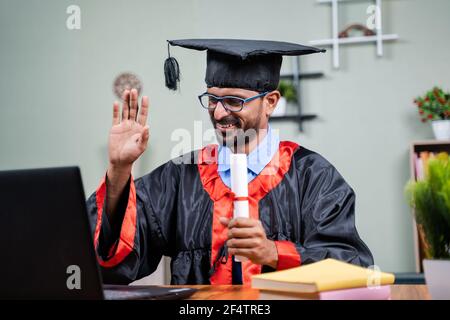 Student attending virtual graduation from laptop by holding certificate in graduation dress from home - concept of virtual celebrations, new normal Stock Photo