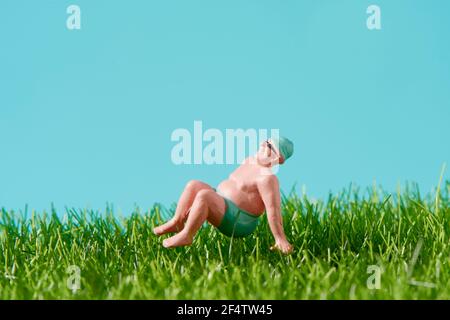 a miniature man, wearing swimsuit and sunglasses, is relaxing sitting on the grass, against a blue background with some blank space on top Stock Photo
