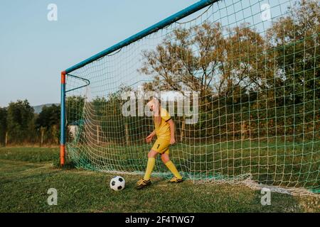 Girl goalkeeper defending goal. Young girl footballer in yellow football dress kicking ball out of net. Stock Photo