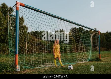 Girl goalkeeper standing at goal on soccer field. Young teenage girl in yellow soccer dress with ball in front of football net. Stock Photo