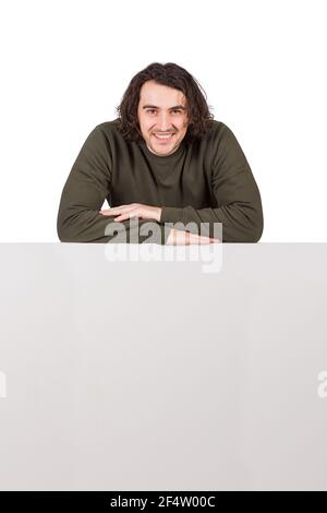 Contented young man, long curly hair style, keeps arms folded leaning on a blank whiteboard, looking confident to camera isolated on white background. Stock Photo