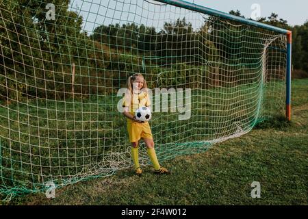 Girl goalkeeper with ball standing at goal on football field. Full length of young teenage girl in yellow soccer dress holding ball in front of soccer Stock Photo