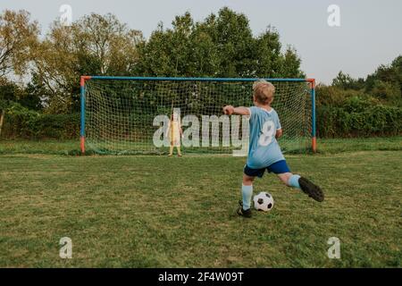 Back view of a child wearing football dress shooting ball in front of goal. Full length of girl goalkeeper standing at goal and soccer player kicking Stock Photo