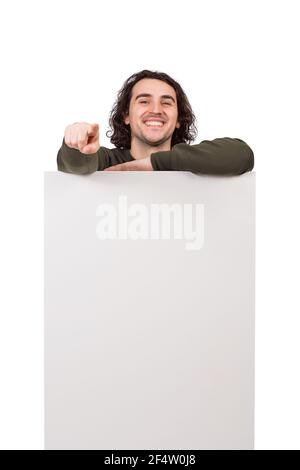 Joyful man standing behind a blank banner for advertising and messages. Happy guy pointing his forefinger showing to camera choosing you. Empty sheet, Stock Photo