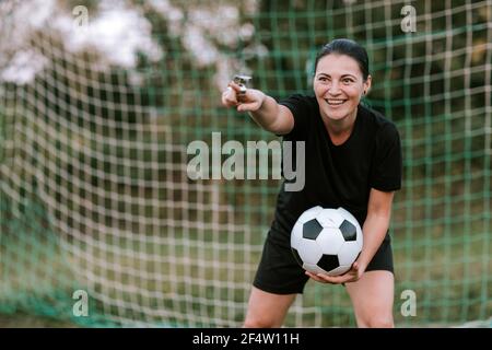 Cheerful female football referee standing at goal on football field. Amateur woman referee in black soccer dress holding ball in front of soccer net. Stock Photo