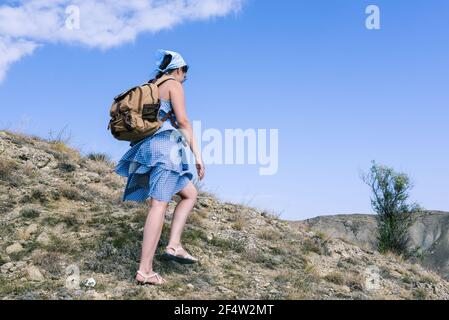 Tourist with a backpack walks along the mountain on a sunny day. Young girl in a blue dress. Active lifestyle and sports. Stock Photo