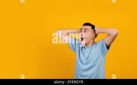 Cool young guy holding his head in shock, screaming OMG and looking aside at empty space on orange studio background Stock Photo