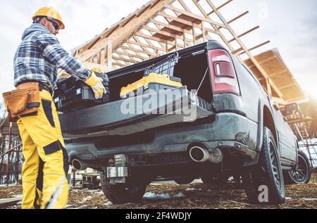 Caucasian Construction Contractor Worker in His 40s Taking Tools Boxes From Bed Section of His Pickup Truck. Newly Constructed Building in Background. Stock Photo