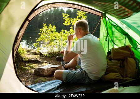 Man making coffee in camping kettle on campfire in forest on shore of a lake, sitting in tent, making a fire, grilling. Happy isolation concept. Stock Photo