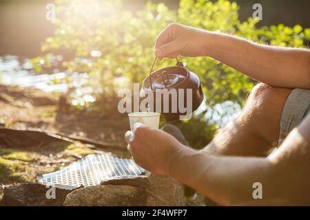 Man making coffee in camping kettle on campfire in forest on shore of a lake, sitting in tent, making a fire, grilling. Happy isolation concept. Stock Photo