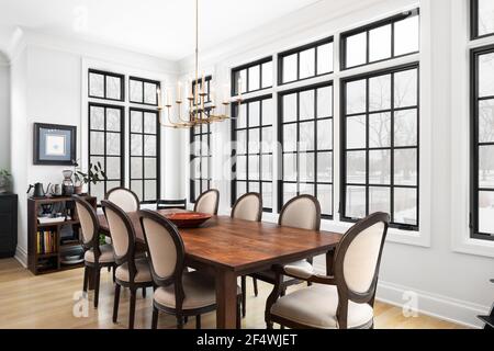 A luxurious dining room with a long wooden table, gold chandelier, black framed windows, hardwood floors, and tall ceilings. Stock Photo