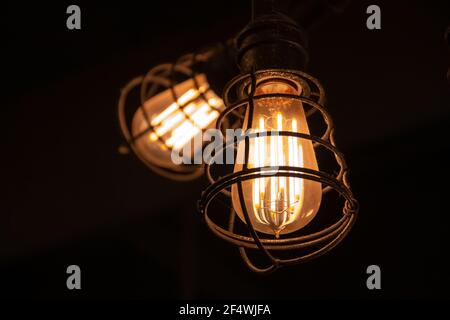 A red-hot factory incandescent light bulb with glowing filaments inside of an iron cage hanging from the ceiling of a pub in London, Ontario, Canada, Stock Photo