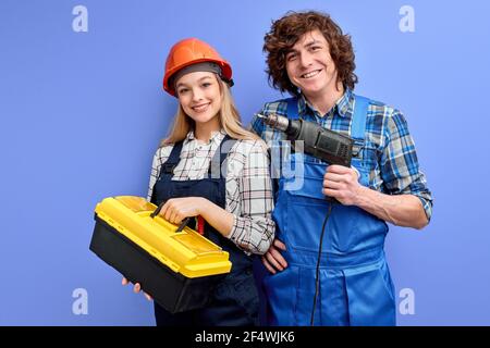team of affable constructors with instruments tools posing isolated on blue background, two builders co-workers standing together looking at camera, d Stock Photo