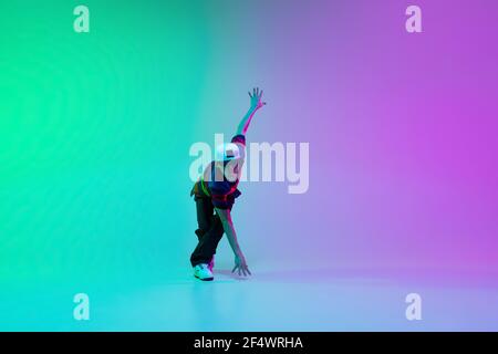 Beautiful sportive boy dancing hip-hop in stylish clothes on colorful gradient background at dance hall in neon light. Stock Photo