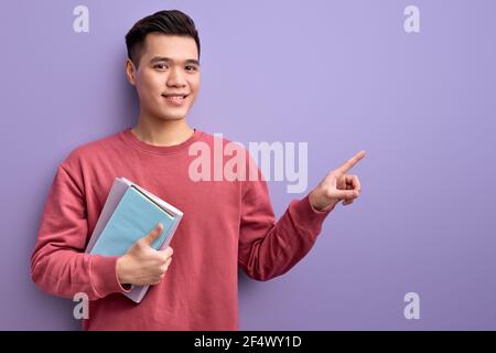 nice asian guy pointing index finger at side holding books in hands, handsome male in casual wear posing, smiling at camera. education concept. copy s Stock Photo