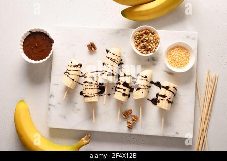 Cooking homemade banana lollipops with different sprinkles on white. Top view. Stock Photo