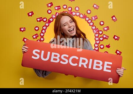 Portrait Of Redhead Female Holding Subscribe Title In Hands, Winking At Camera Happily, Isolated On Yellow Background Stock Photo