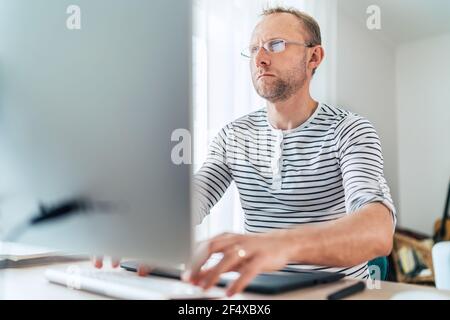 A middle-aged man in eyeglasses typing a text using a modern keyboard computer in his living room. Writing or Distance or freelance work on worldwide Stock Photo