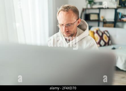 A middle-aged man in eyeglasses using a modern computer in his living room. Writing or Distance or freelance work on worldwide quarantine time concept Stock Photo