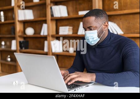 Focused African-American wearing medical mask working with a laptop in contemporary office space with bookshelves on the background, social distancing and protective measures for office employees Stock Photo