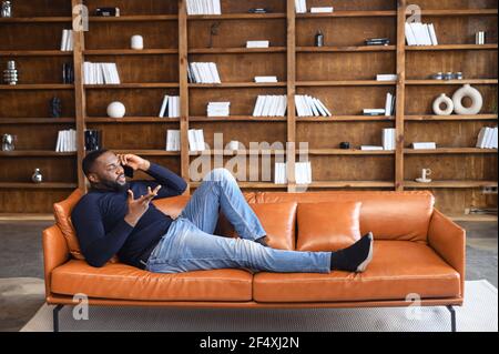 Full length an African-American young man lying on the coack and talking over the smartphone, confident multiracial male freelancer has phone conversation working from home, book shelves on background Stock Photo