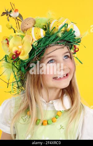 Happy girl having fun Easter egg hunt, wearing straw nest hairstyle made of spring flowers, Easter eggs and feathers. The child looks to the side smil Stock Photo
