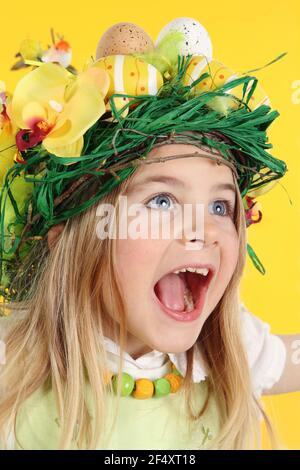 Young happy girl wearing straw headdress made of spring flowers, Easter eggs and feathers. The child with the big eyes is surprised by happiness. Art Stock Photo