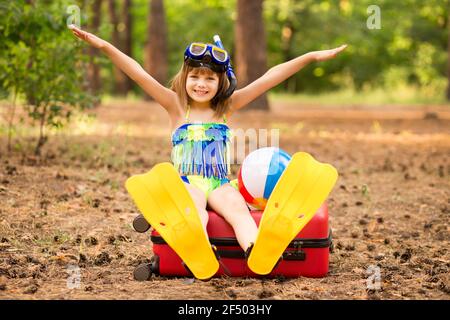 Little child girl wear swimsuit sitting on suitcase in summer forest with hands up, show plane. Dream journey. Vacation concept Stock Photo