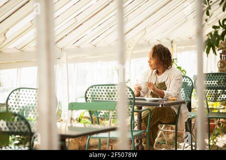 Female garden shop owner drinking tea at table in greenhouse Stock Photo