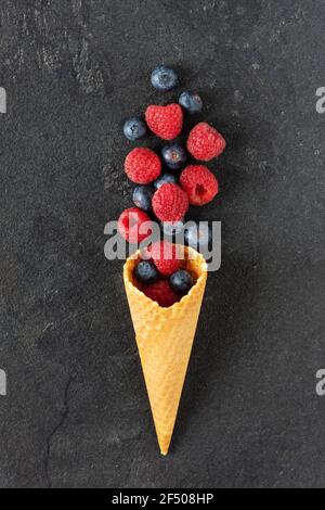Mix of blueberry and raspberry berries in a waffle ice cream cone on black background top view. Creative composition with wild summer berries. Stock Photo