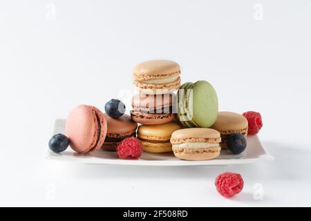 Multi colored french macaroons and berries such as raspberry and blueberry on a plate over white background. French meringue cookie macarons and berri Stock Photo