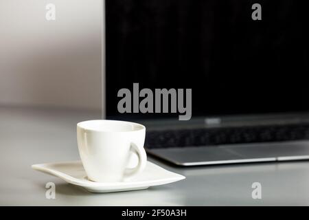 Opened laptop and coffee cup on saucer in the foreground on the grey table and white background. Remote workplace, home office. Online communication. Stock Photo