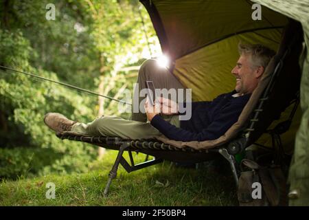 Man in camping lounge chair fishing and using digital tablet Stock Photo