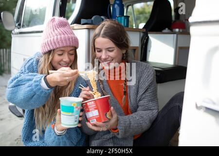 Young women friends sharing instant noodles at camper van doorway Stock Photo