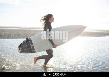 Happy young female surfer running with surfboard in sunny ocean surf Stock Photo